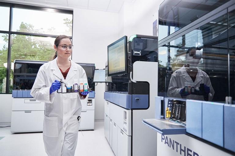 Kvinnlig laboratorietekniker bär en bricka med prover som ska sättas in i Panther-systemet