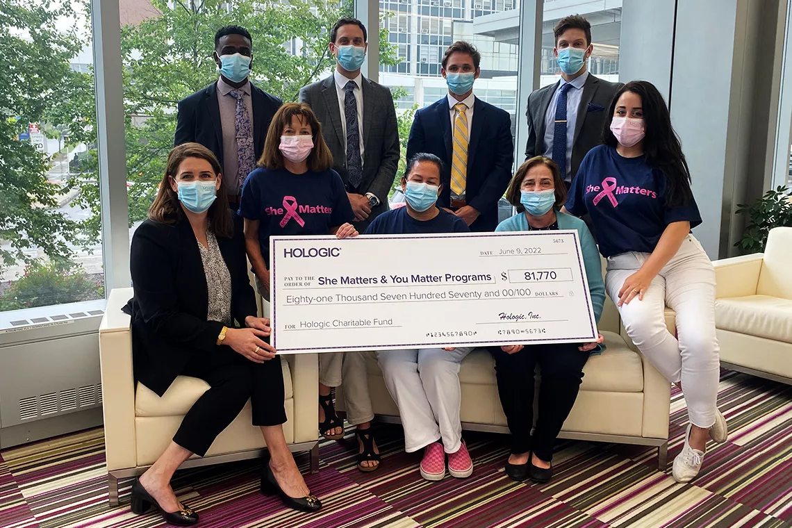 Gruppbild på personer med munskydd som håller en stor kampanjcheck till stöd för programmen She Matters och You Matter